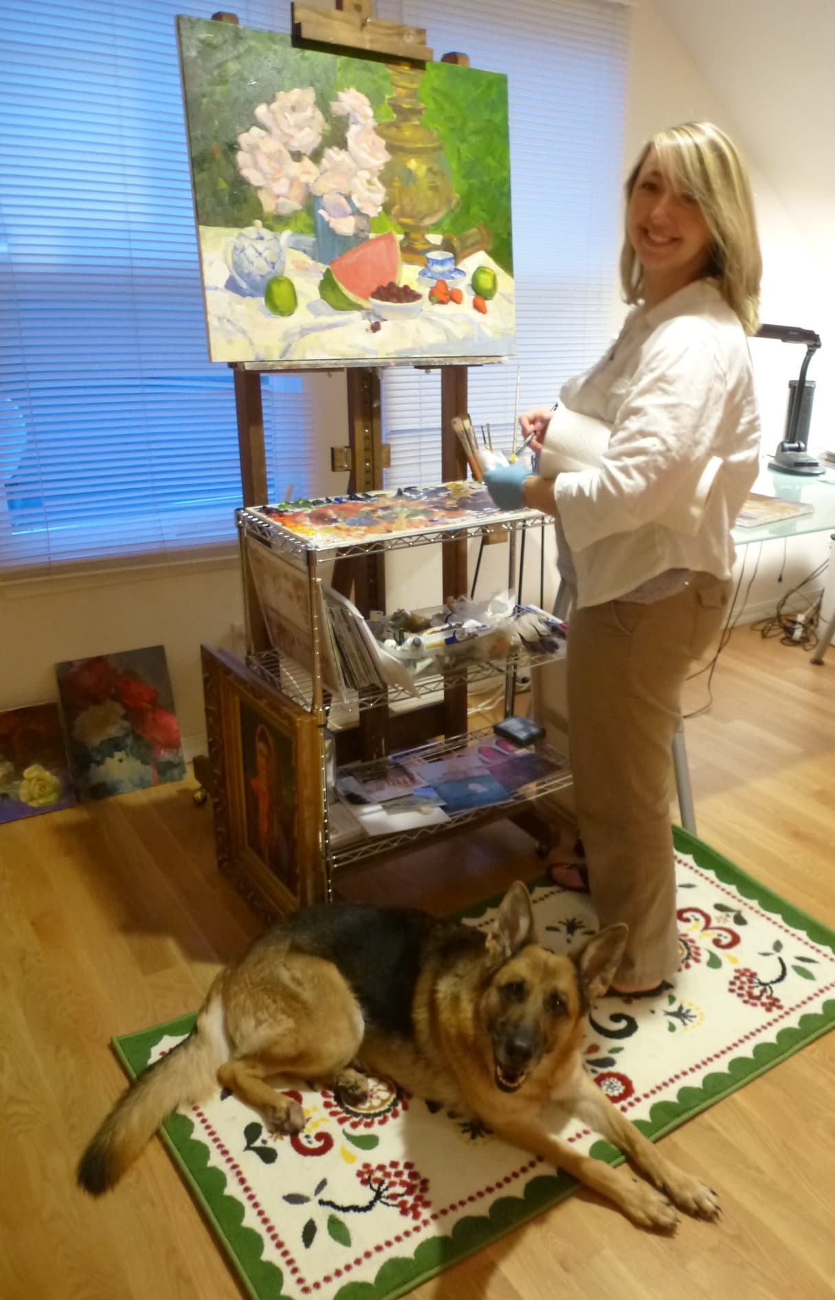 Elena Balekha standing at her easel with a palette in her hand, painting a still life. Her German Shepherd Roxy lies on the rug in front of her.